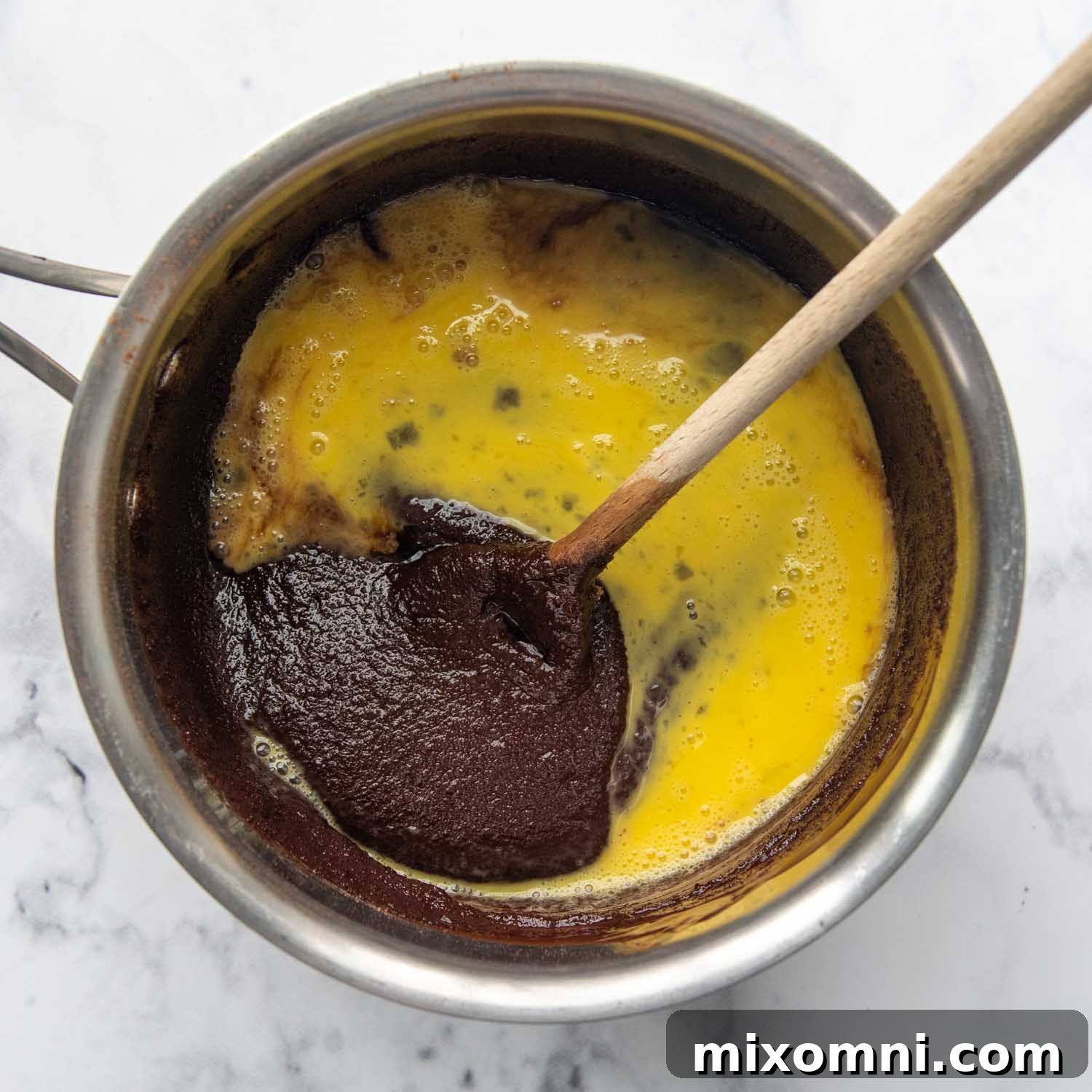 Two large, lightly beaten eggs being added to the cooled chocolate batter in the saucepan.