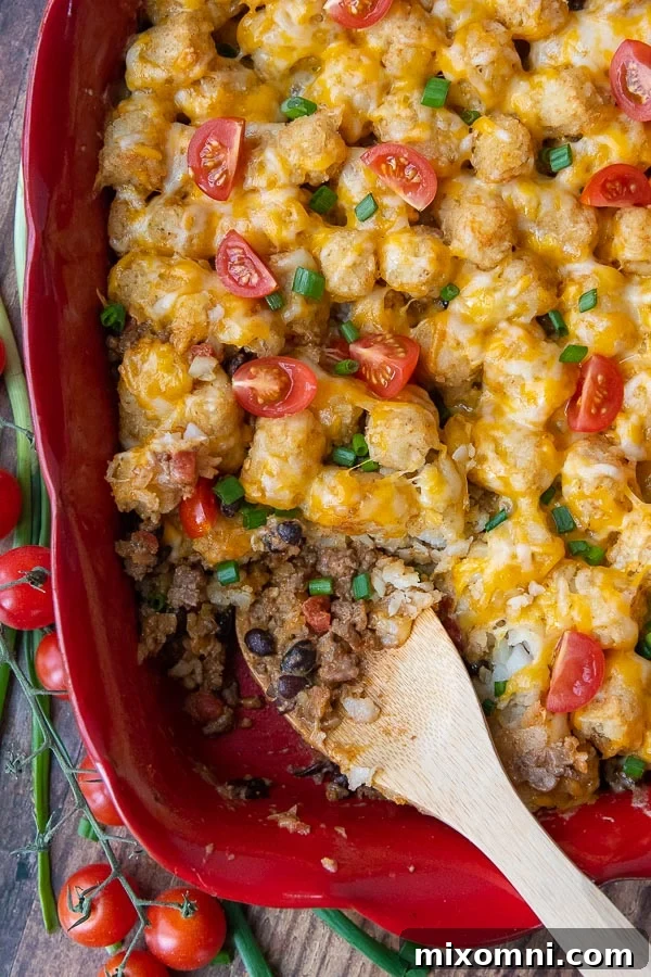 Overhead shot of a Mexican Tater Tot Casserole in a baking dish with a scoop taken out, revealing layers of seasoned ground beef, black beans, and melted cheese under crispy tater tots. Garnished with cilantro.
