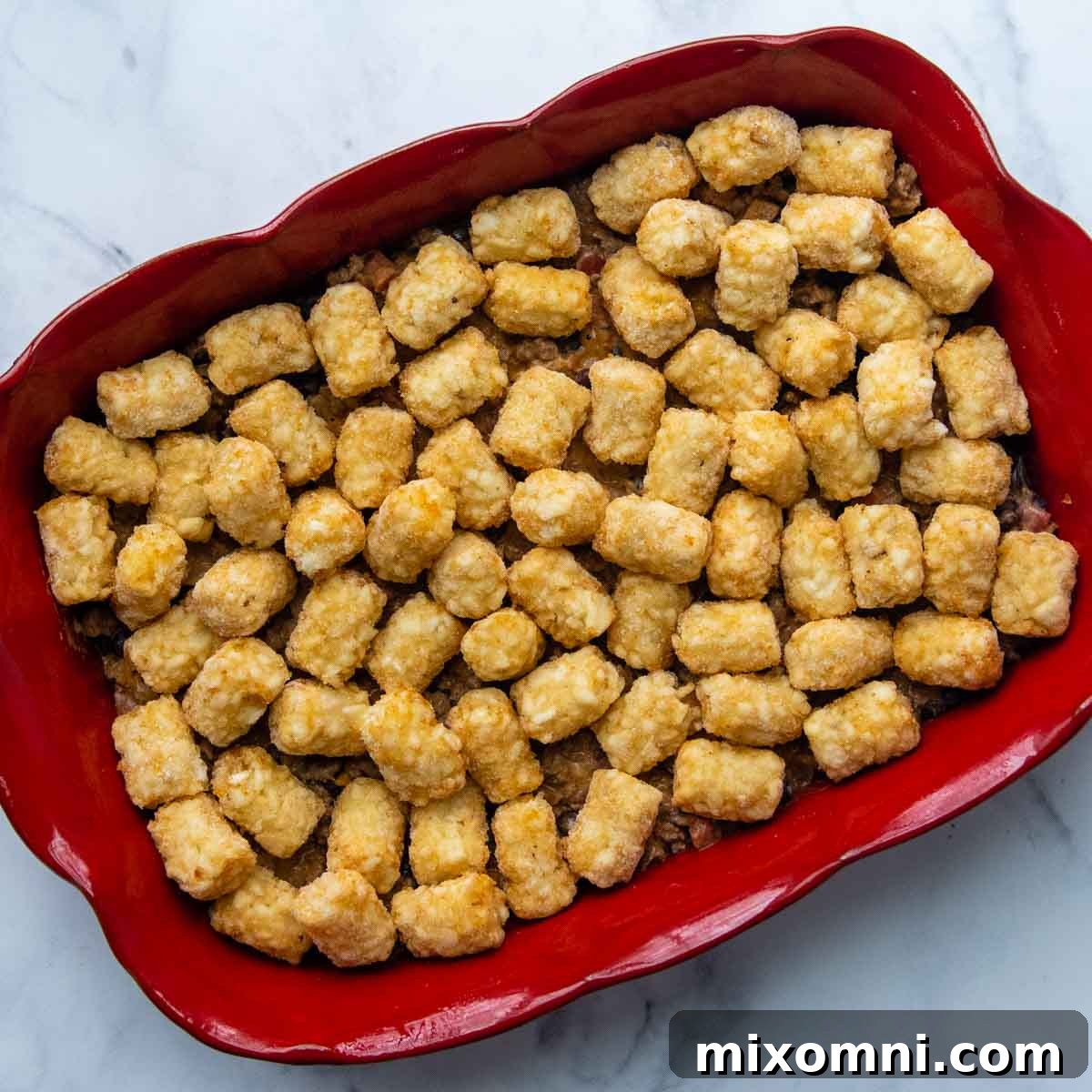 Unbaked frozen tater tots arranged in a single layer on top of the beef mixture in a casserole dish.