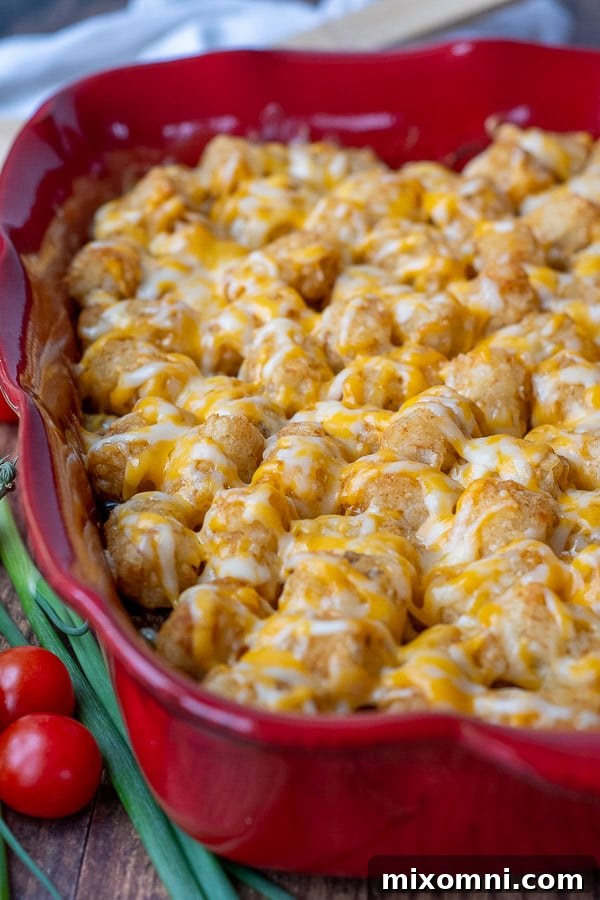 Close-up picture of Mexican tater tot casserole in a red baking dish, showing the melted cheese, crispy tater tots, and bubbling beef mixture underneath.