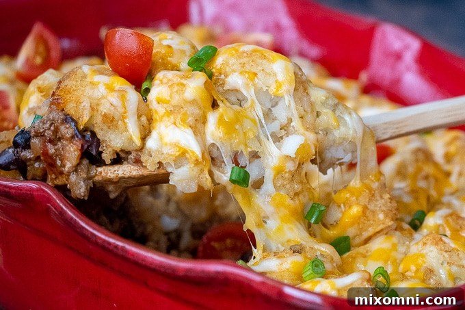 A wooden spoon lifting a scoop of Mexican tater tot casserole from a red baking dish, showing the crispy tots, melted cheese, and hearty beef mixture.