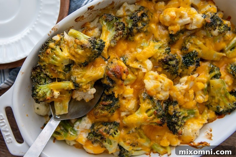 A large serving spoon lifts a generous portion of cheesy broccoli and cauliflower casserole from a baking dish.