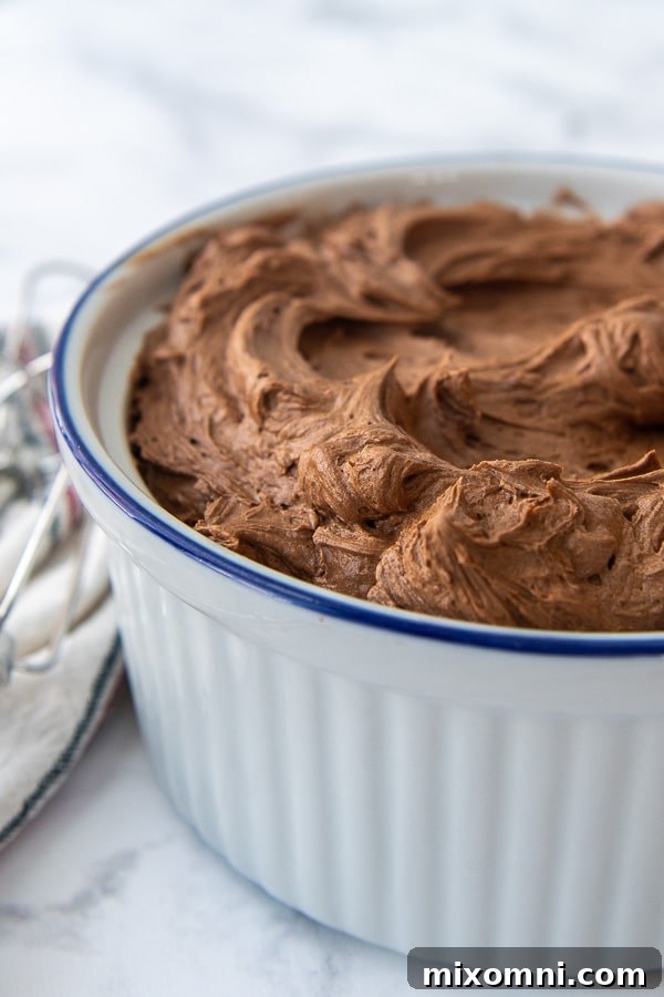 Close-up side angle shot of super fluffy chocolate buttercream frosting in a pristine white bowl, with mixing beaters resting nearby.