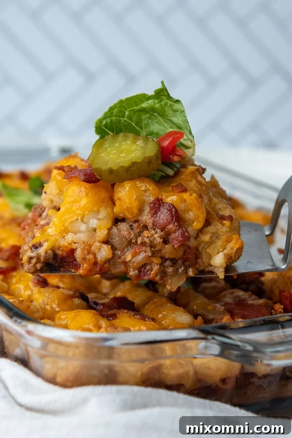A spatula lifting a portion of bacon cheeseburger casserole from a baking dish, showcasing melted cheese, crispy tater tots, and a rich beef base.
