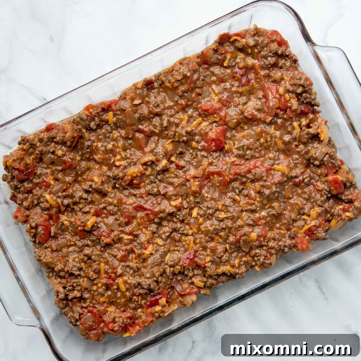 The seasoned ground beef and bacon mixture spread evenly across the bottom of a glass baking dish, forming the base of the casserole.