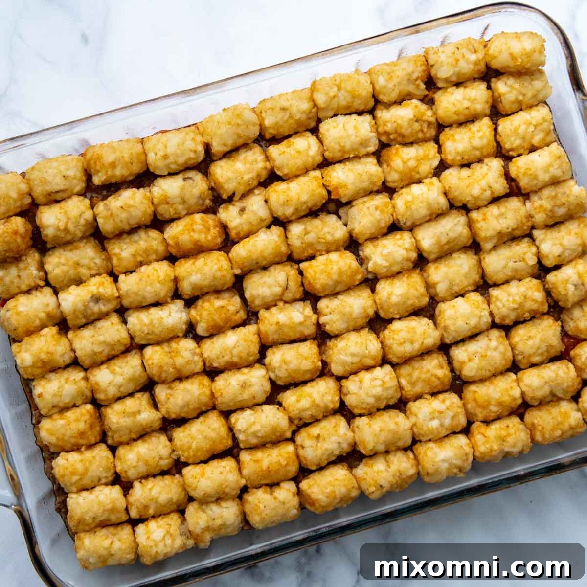 Frozen tater tots arranged in neat rows on top of the ground beef mixture in a baking dish, ready for baking.