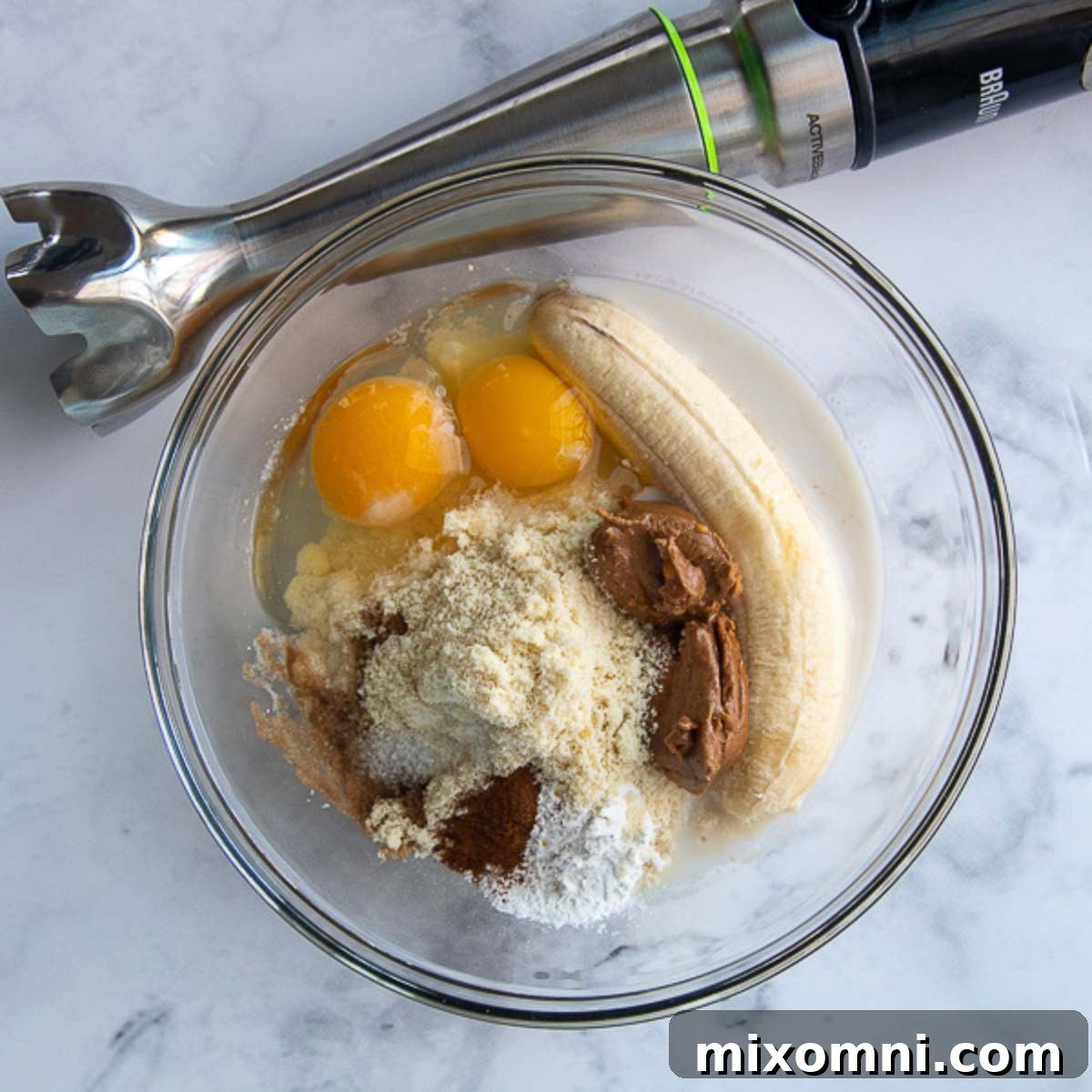 All the ingredients for the pancakes are shown in a glass bowl, with an immersion blender poised beside it, ready to mix. The scene suggests fresh, wholesome ingredients waiting to be combined.