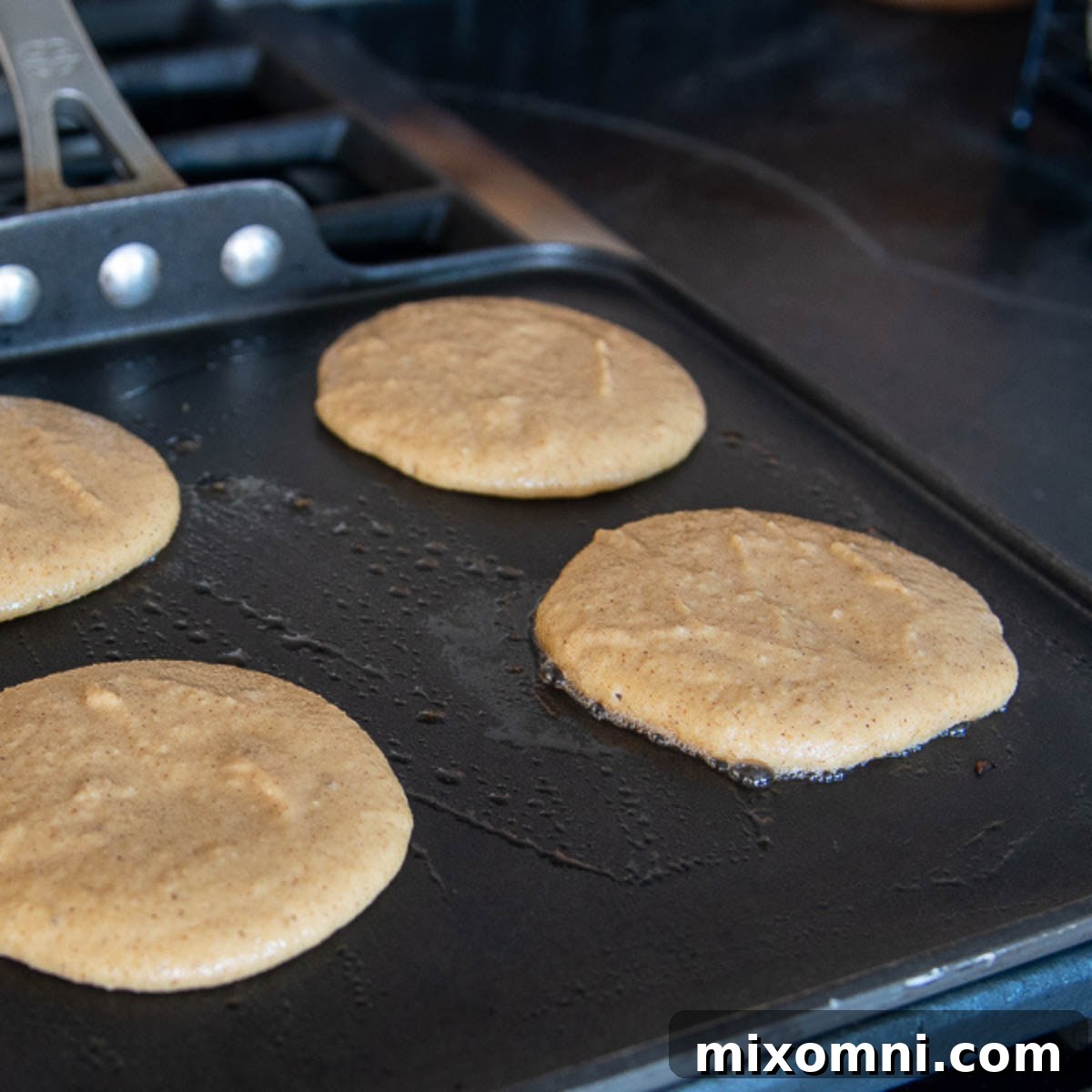 Several pancakes are cooking on a griddle, showing the golden-brown edges and gentle bubbling that indicate they are almost ready to be flipped. The heat is visible, suggesting a moderate temperature for perfect cooking.