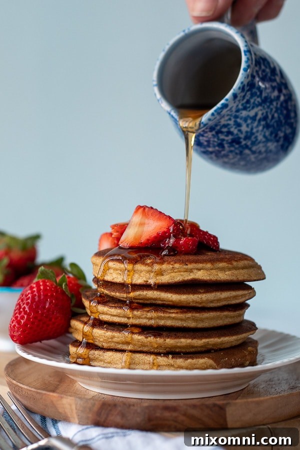 A generous stream of golden maple syrup being poured over a tall, inviting stack of warm almond flour pancakes. The syrup glistens, highlighting the soft texture of the pancakes, suggesting a truly indulgent breakfast.