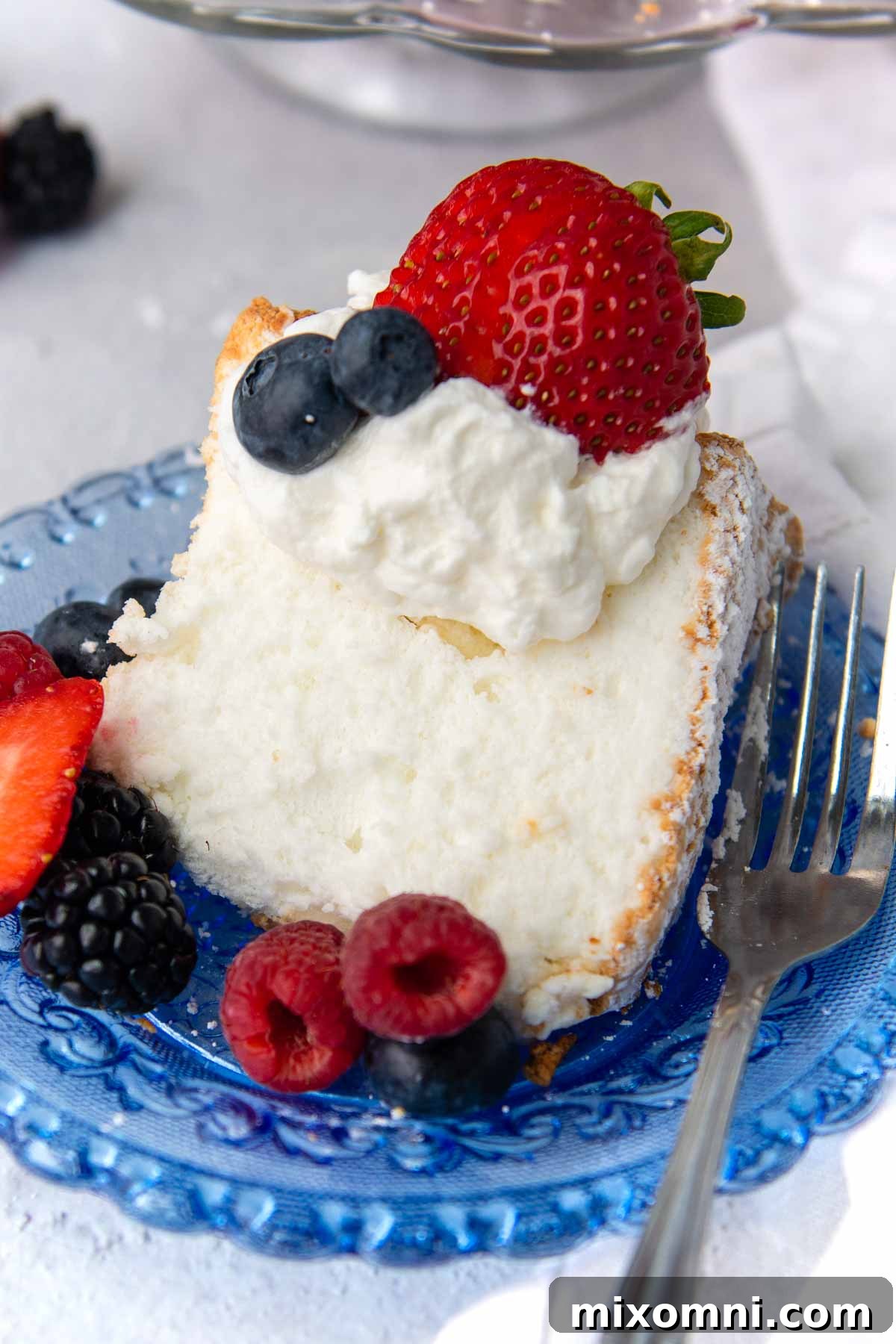 A delicate slice of angel food cake on a blue plate, garnished with whipped cream and fresh fruit.