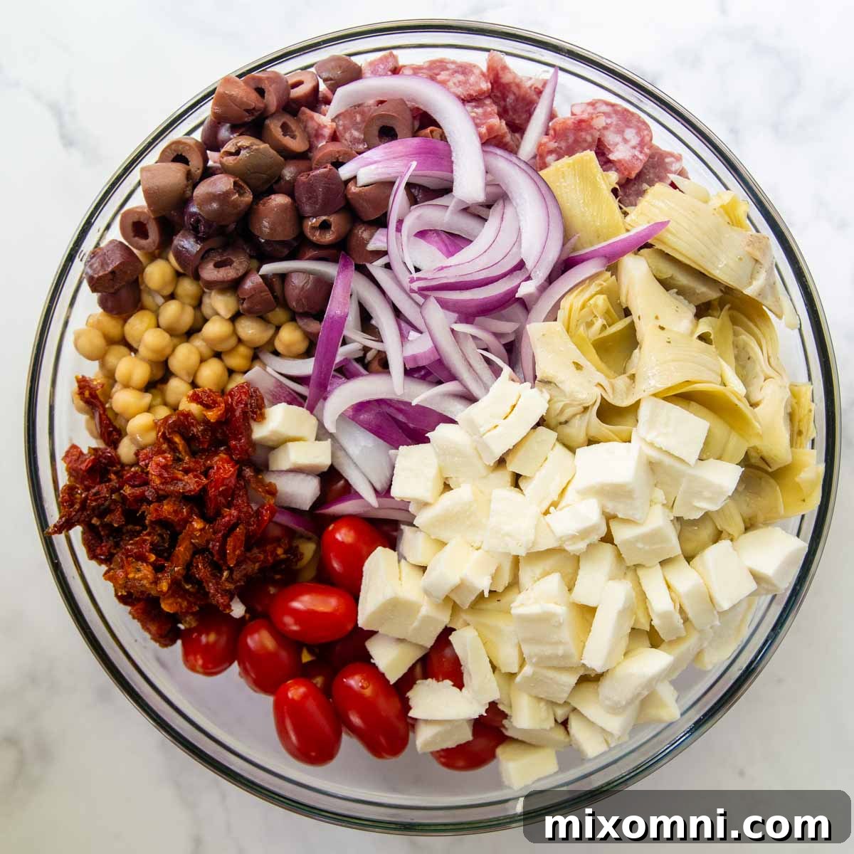 A large mixing bowl filled with cooked gluten-free pasta and a colorful array of ingredients like tomatoes, olives, artichoke hearts, and salami, ready to be combined.