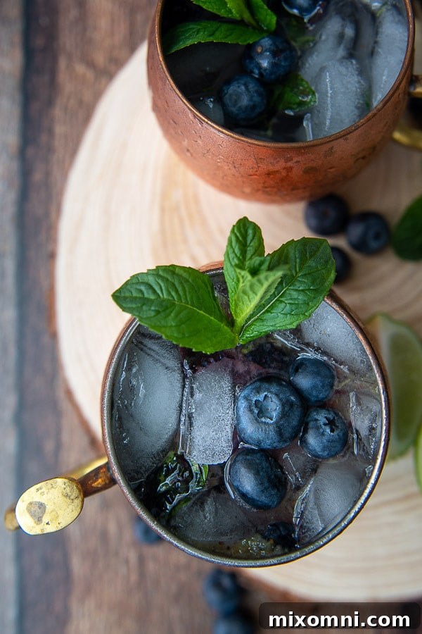 Another appealing overhead shot of two flavored Moscow Mules, beautifully presented on a wooden circle board, garnished with fresh blueberries and mint, emphasizing their delightful presentation.