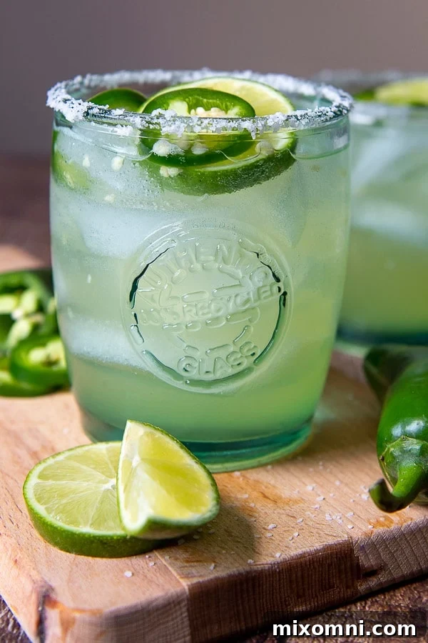 A vibrant spicy margarita garnished with lime and jalapeño slices, served on a wooden cutting board amidst fresh limes and peppers.