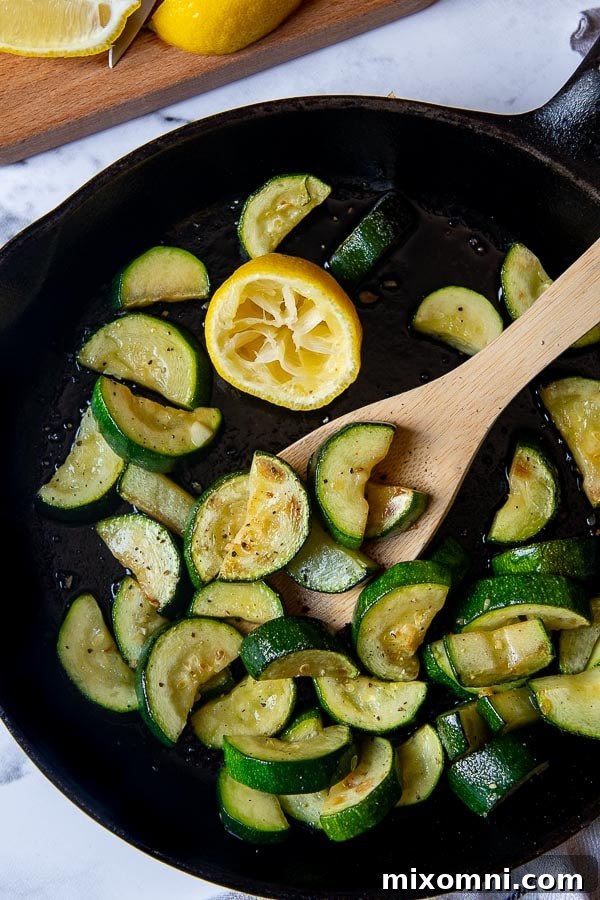 Overhead shot of freshly sauteed zucchini and squash in a black cast iron skillet, garnished with fresh herbs and a halved lemon, ready to be served.