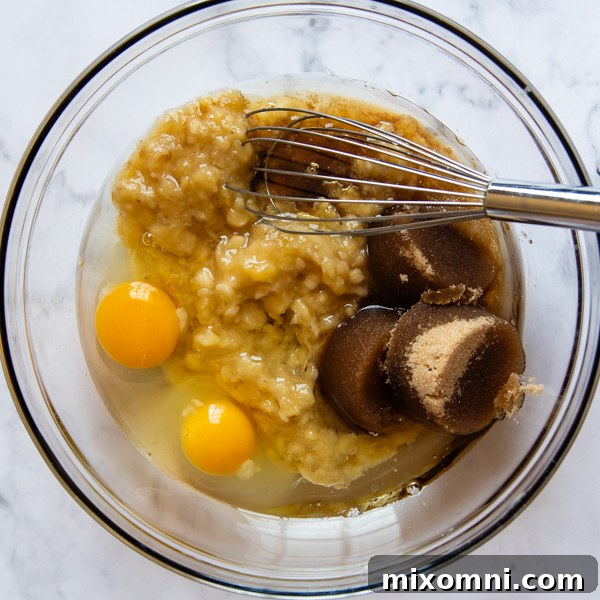 A large mixing bowl containing mashed bananas, eggs, oil, and brown sugar being vigorously whisked together to combine the wet ingredients for gluten-free banana bread.