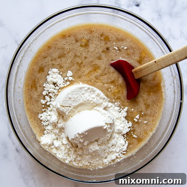 A mixing bowl filled with gluten-free banana bread batter, with flour being stirred in to combine all the ingredients.