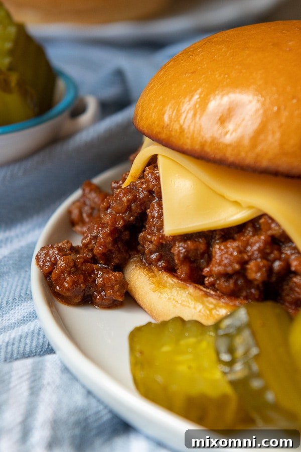 Close-up of sloppy joe meat dripping out of a toasted bun, with pickles in the foreground, ready to be enjoyed.