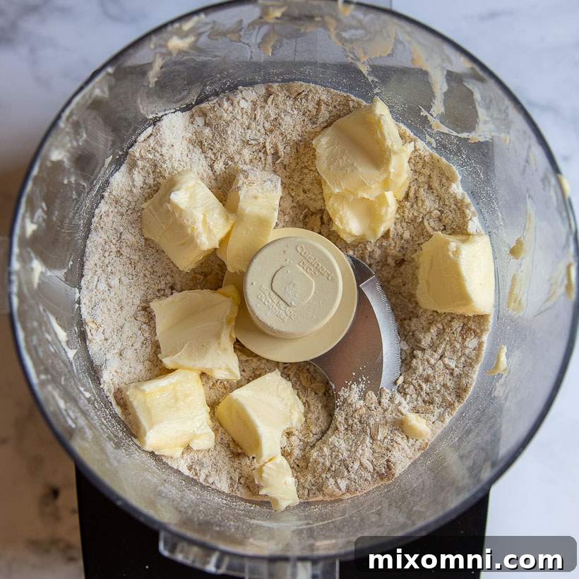 Cubed, softened butter pieces are about to be added to the dry oat topping mixture in a food processor, signaling the next step in creating the crisp.