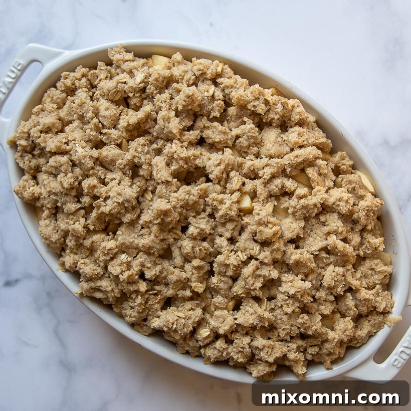 An unbaked gluten-free apple crisp, nestled in a white oval baking dish, covered with a thick, clumpy oat topping, awaiting its golden transformation in the oven.