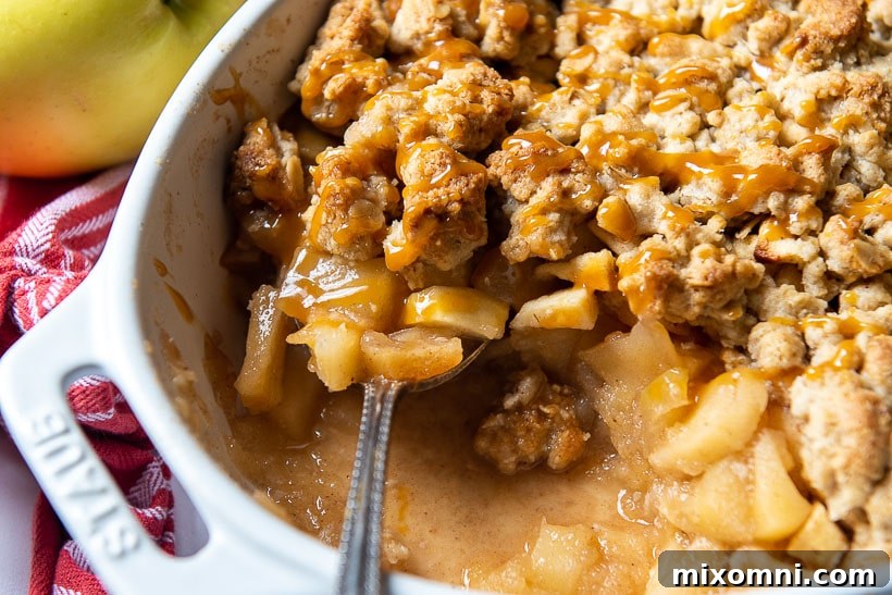 A spoon delicately lifting out a generous portion of warm gluten-free apple crisp, showcasing the golden, crunchy topping and gooey apple filling, with a rich caramel sauce cascading over it.