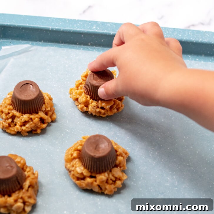 A child's hand gently placing a peanut butter cup onto a newly formed crispy rice cookie, showcasing kid-friendly involvement.