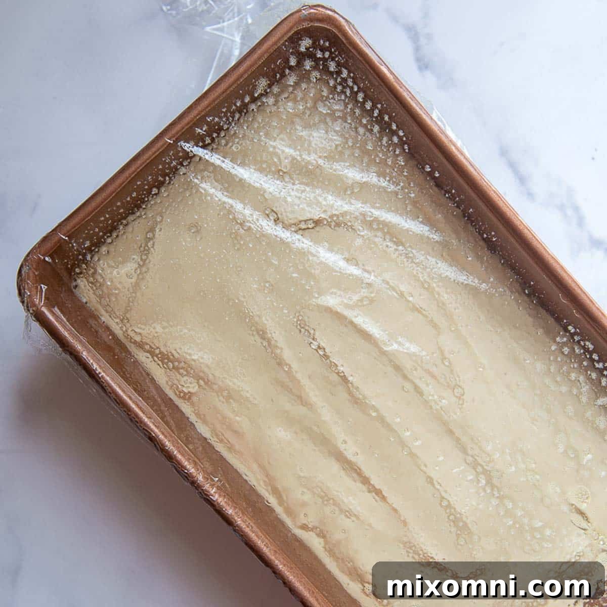 A bread pan containing rising gluten-free dough, covered with plastic wrap, in a warm place.
