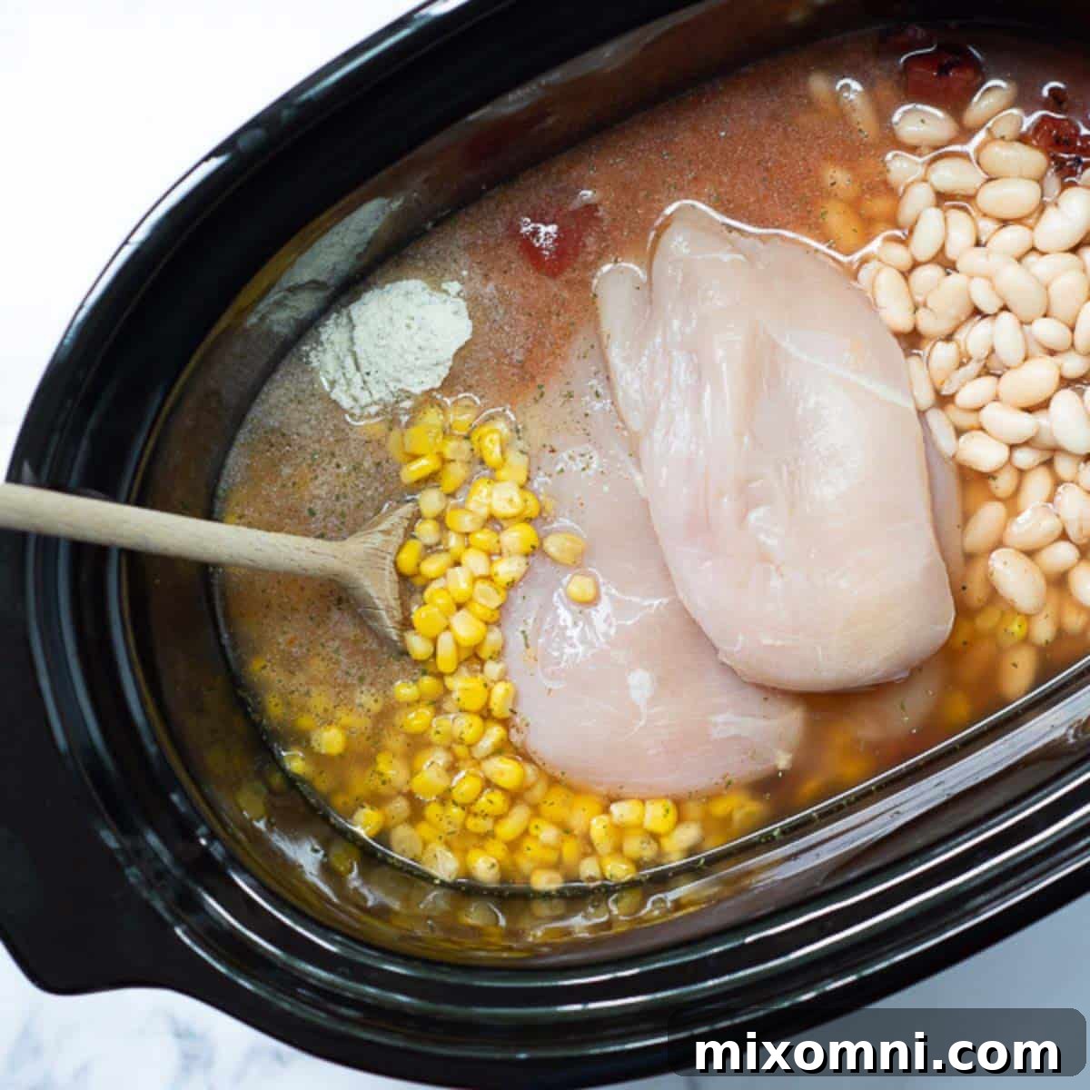 All the raw ingredients for creamy buffalo chicken chili, including chicken, beans, tomatoes, and spices, are assembled in a slow cooker, ready for cooking.