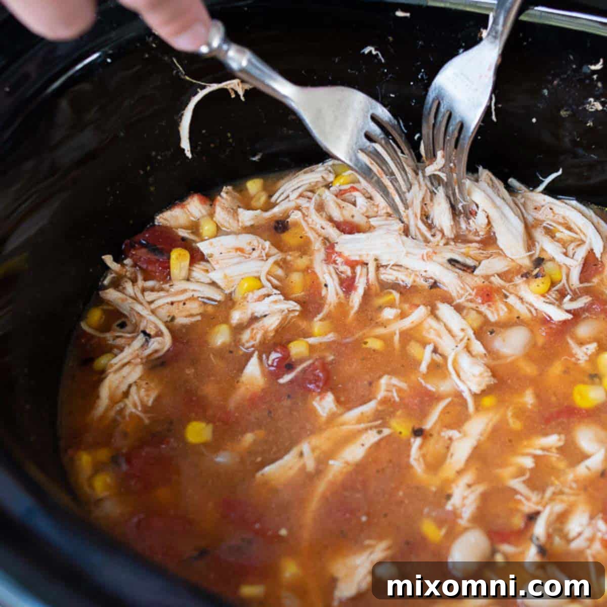 A pair of forks actively shredding tender, cooked chicken breasts directly in the slow cooker, surrounded by the chili's rich, creamy base.