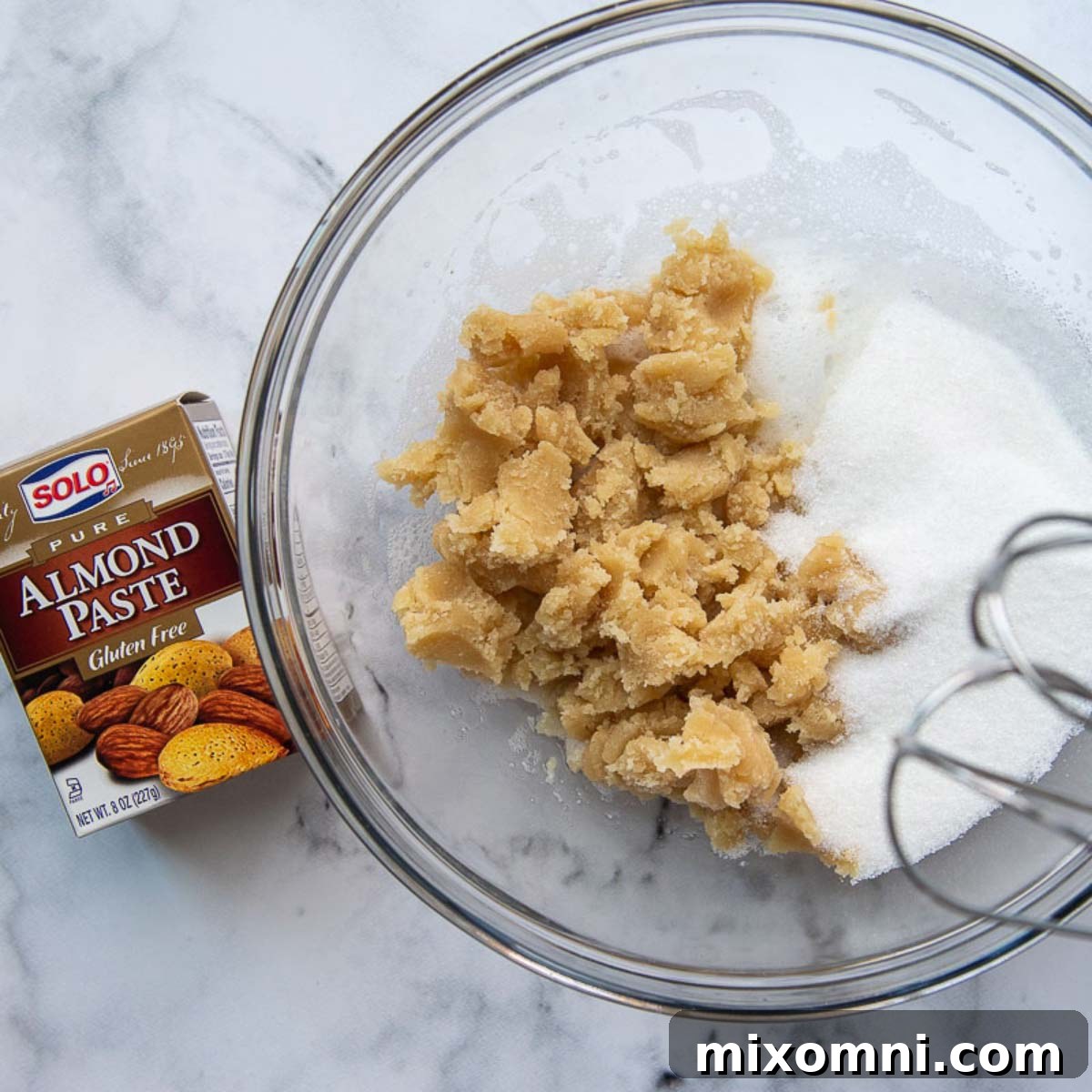 All the essential ingredients for almond cookies, including chunks of almond paste, sugar, and almond flour, gathered in a large glass bowl, ready for mixing.