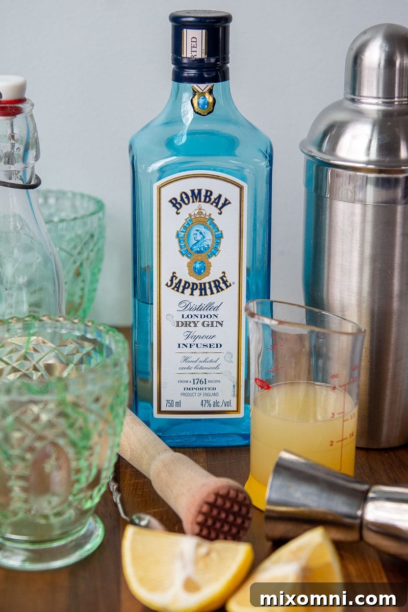 A perfectly arranged selection of ingredients for making a gin smash cocktail, including fresh basil leaves, whole lemons, a bottle of gin, and a container of simple syrup, all laid out on a rustic surface.