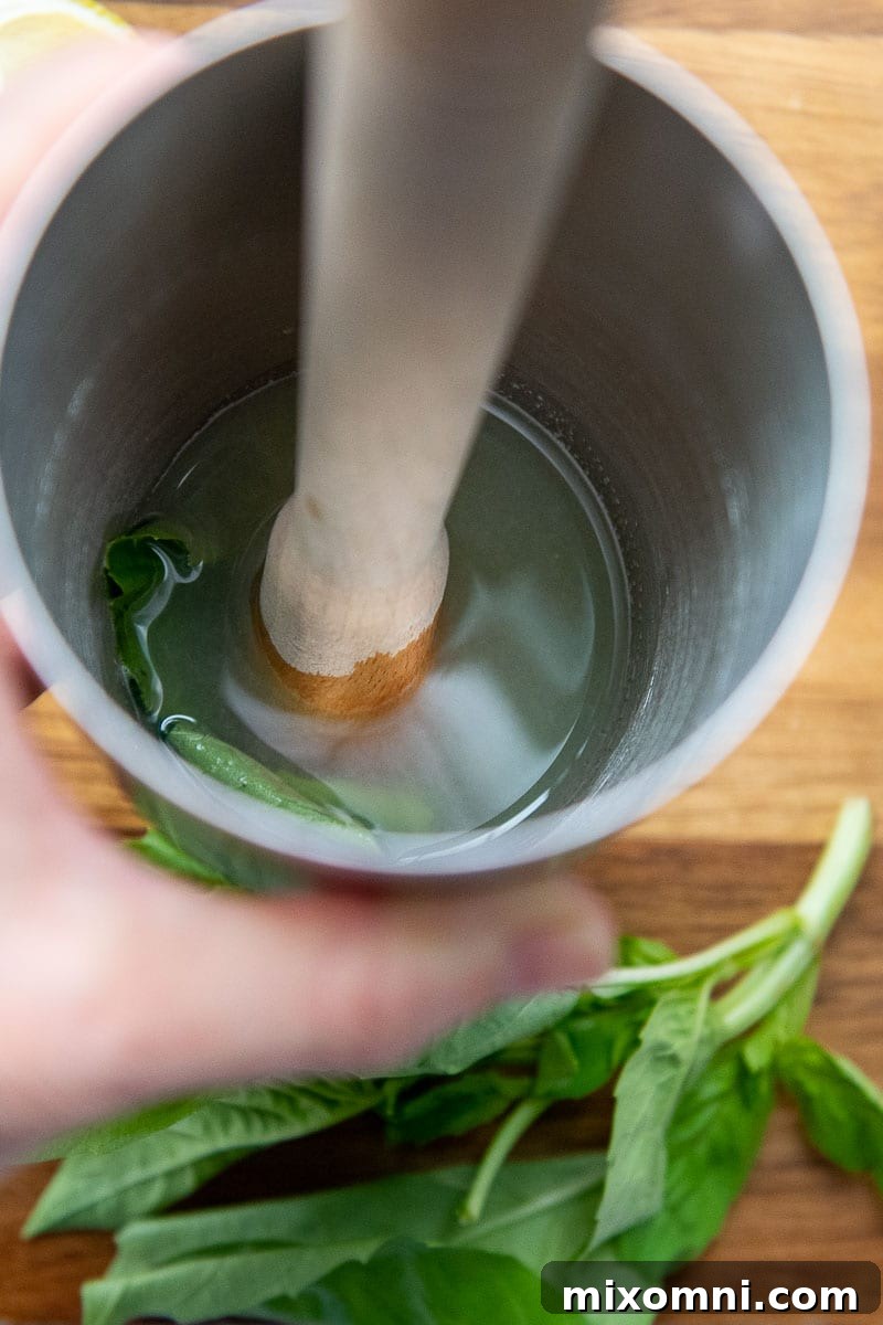 A muddler gently pressing fresh basil leaves, simple syrup, and lemon juice together inside a cocktail shaker, illustrating the key step of releasing the basil's flavors.