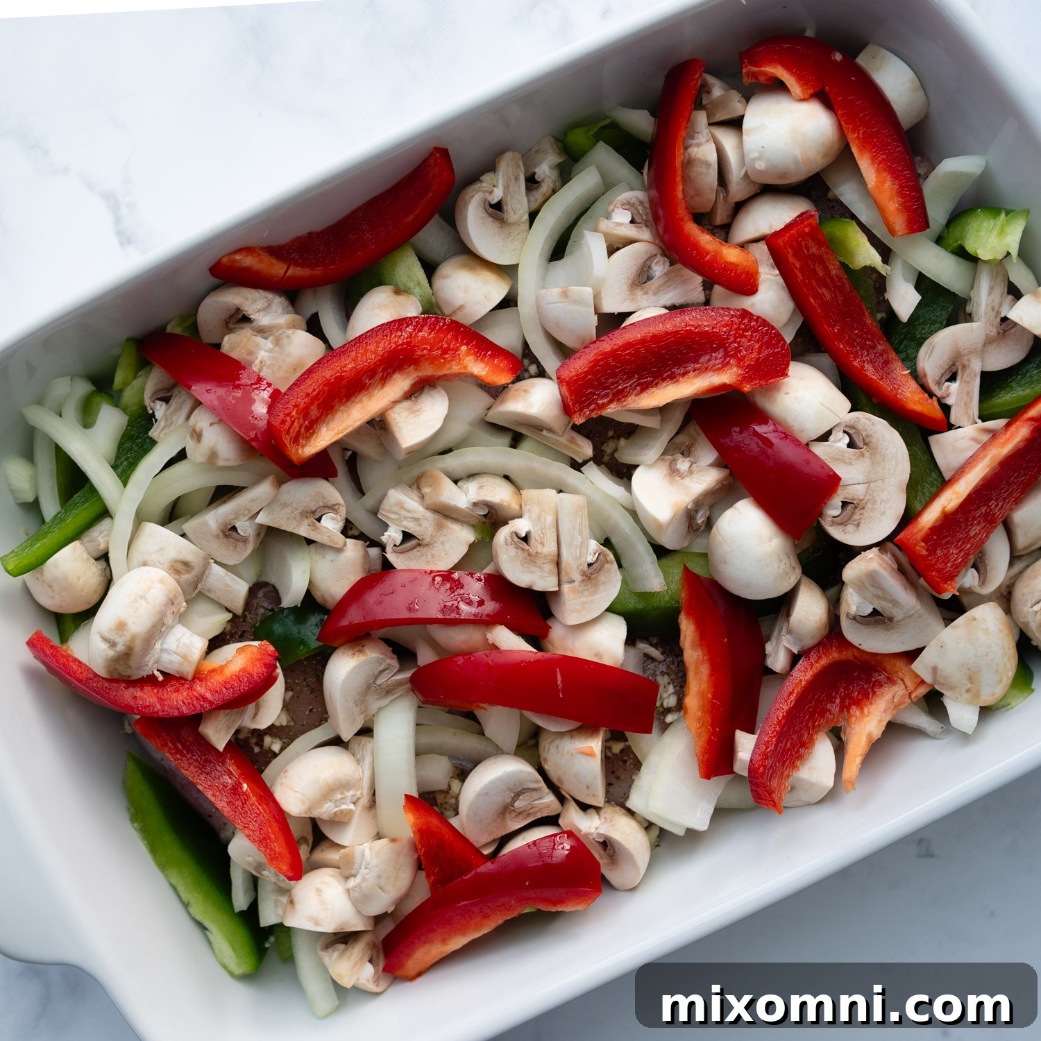 sliced mushrooms, onions, and bell peppers layered over the seasoned raw chicken in a baking dish.