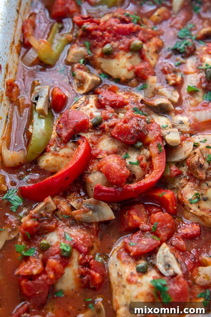close up of baked chicken with peppers, mushrooms, and onions in a rich tomato sauce, garnished with fresh herbs.
