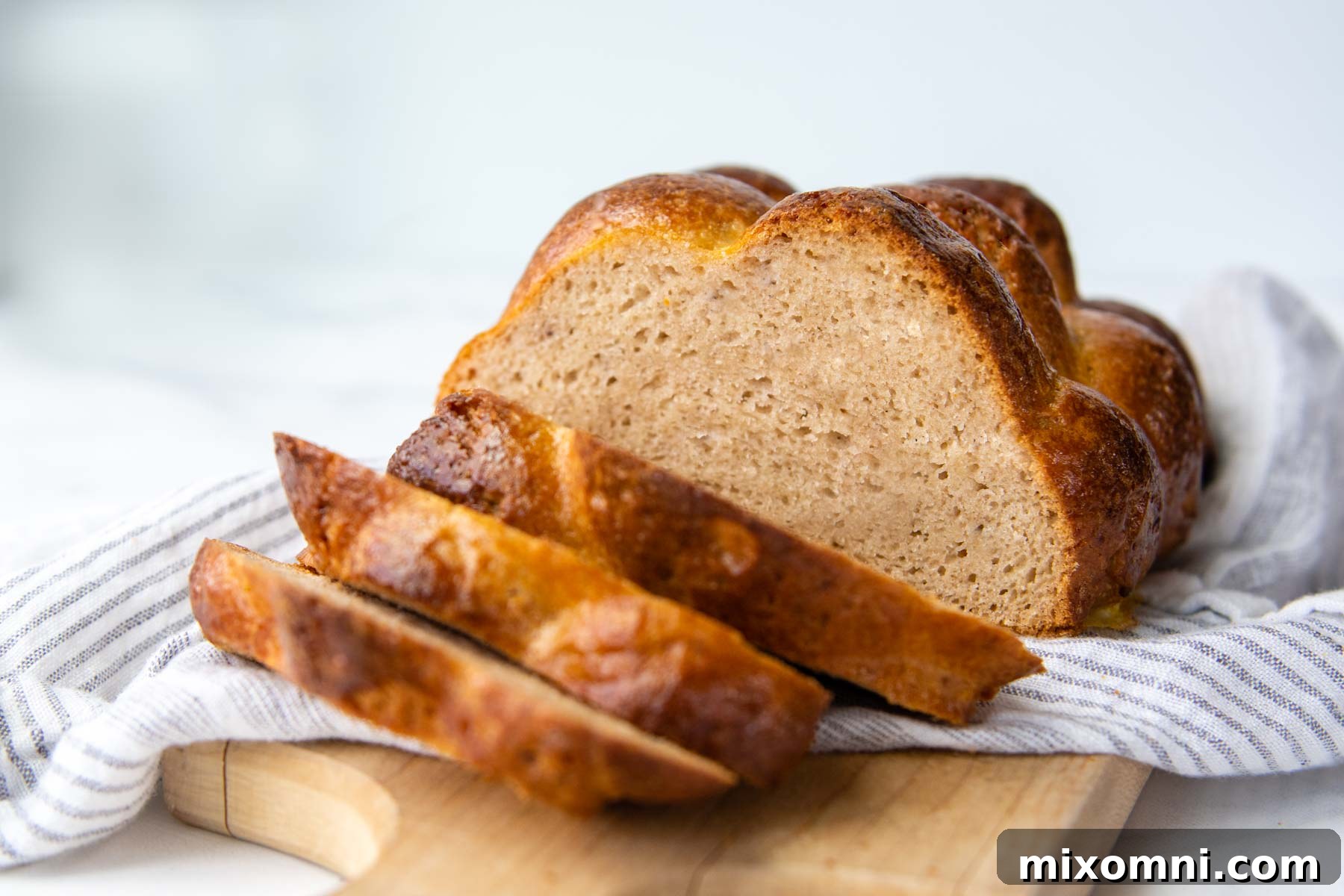 A half-sliced loaf of golden gluten-free challah bread on a cutting board, resting on a white linen, ready to be served.