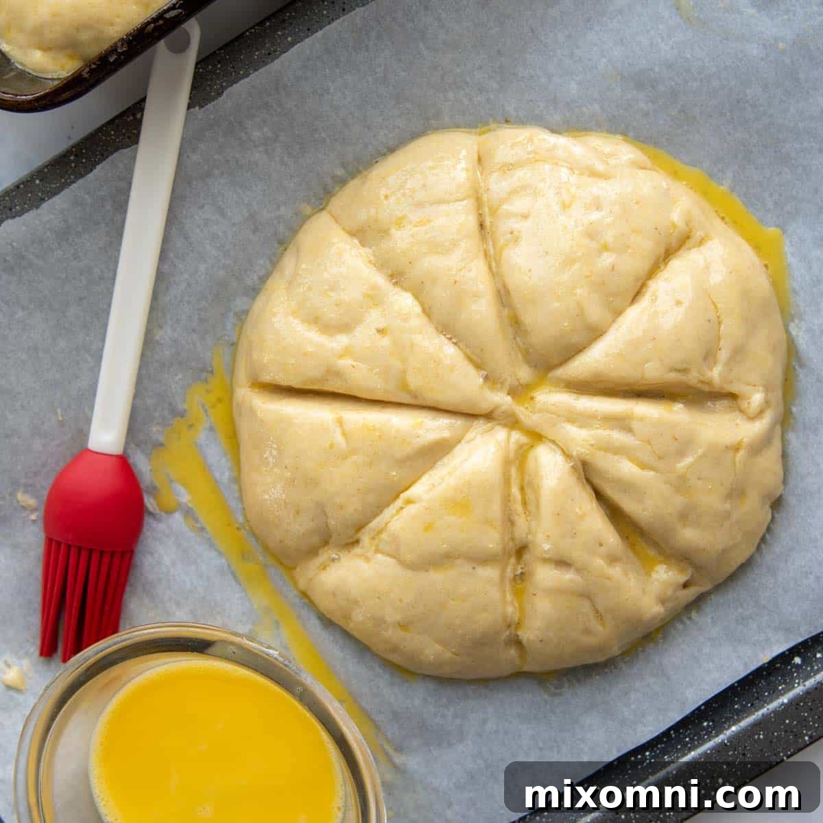 A round challah loaf on a baking sheet, scored with a star pattern before baking, next to an egg wash brush.
