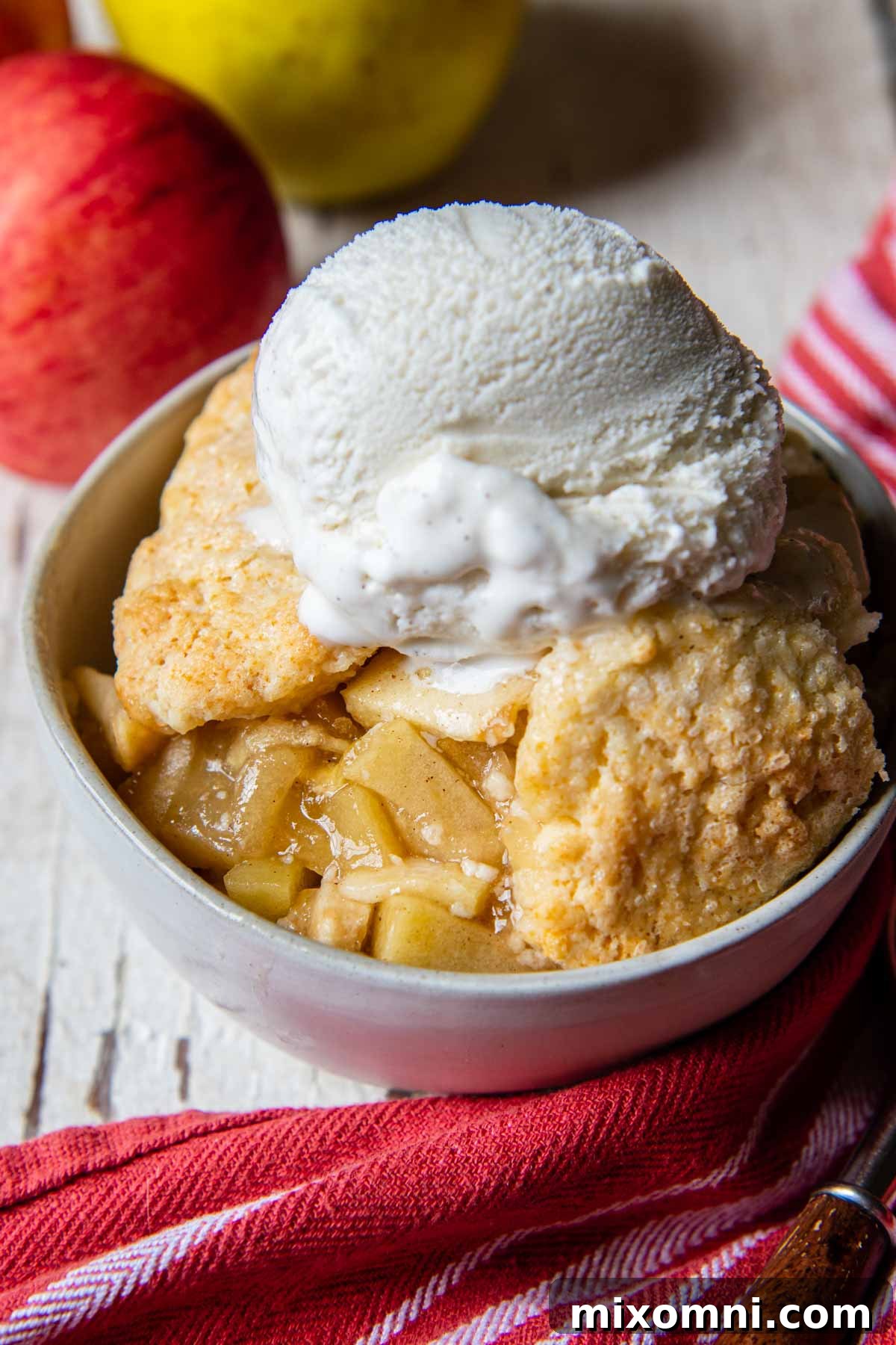 A delectable serving of golden-brown gluten-free apple cobbler in a white bowl, with fresh apples artfully arranged in the background, suggesting wholesome ingredients.