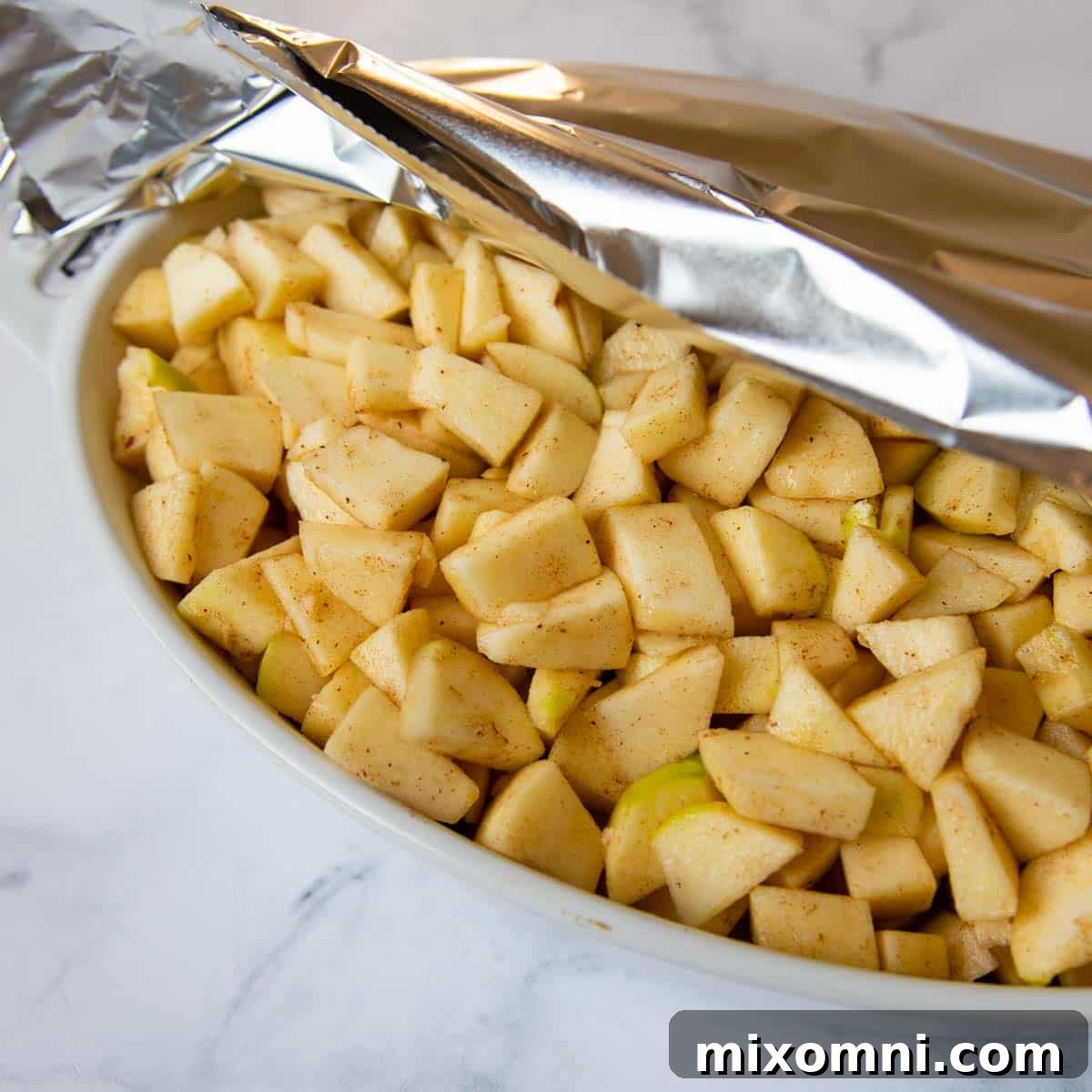 A baking dish filled with cinnamon-spiced apples, partially covered with aluminum foil and ready to be baked, showcasing the pre-baking step for a perfectly tender filling.