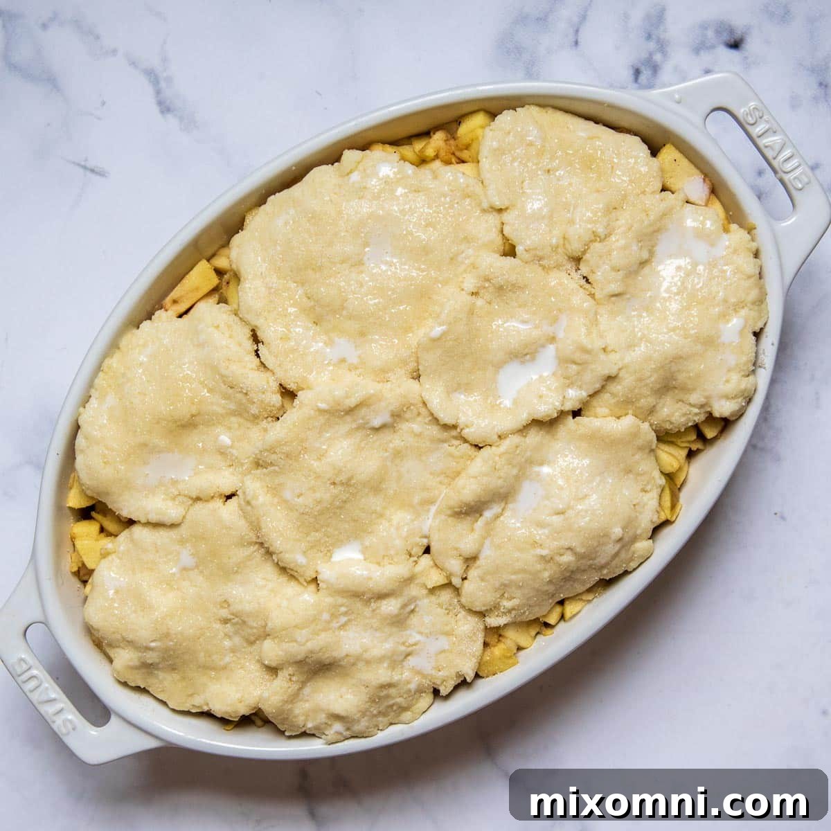 Delicate, flat rounds of gluten-free biscuit dough artfully placed atop the bubbling apple filling in a baking dish, ready for their final bake into a golden cobbler.