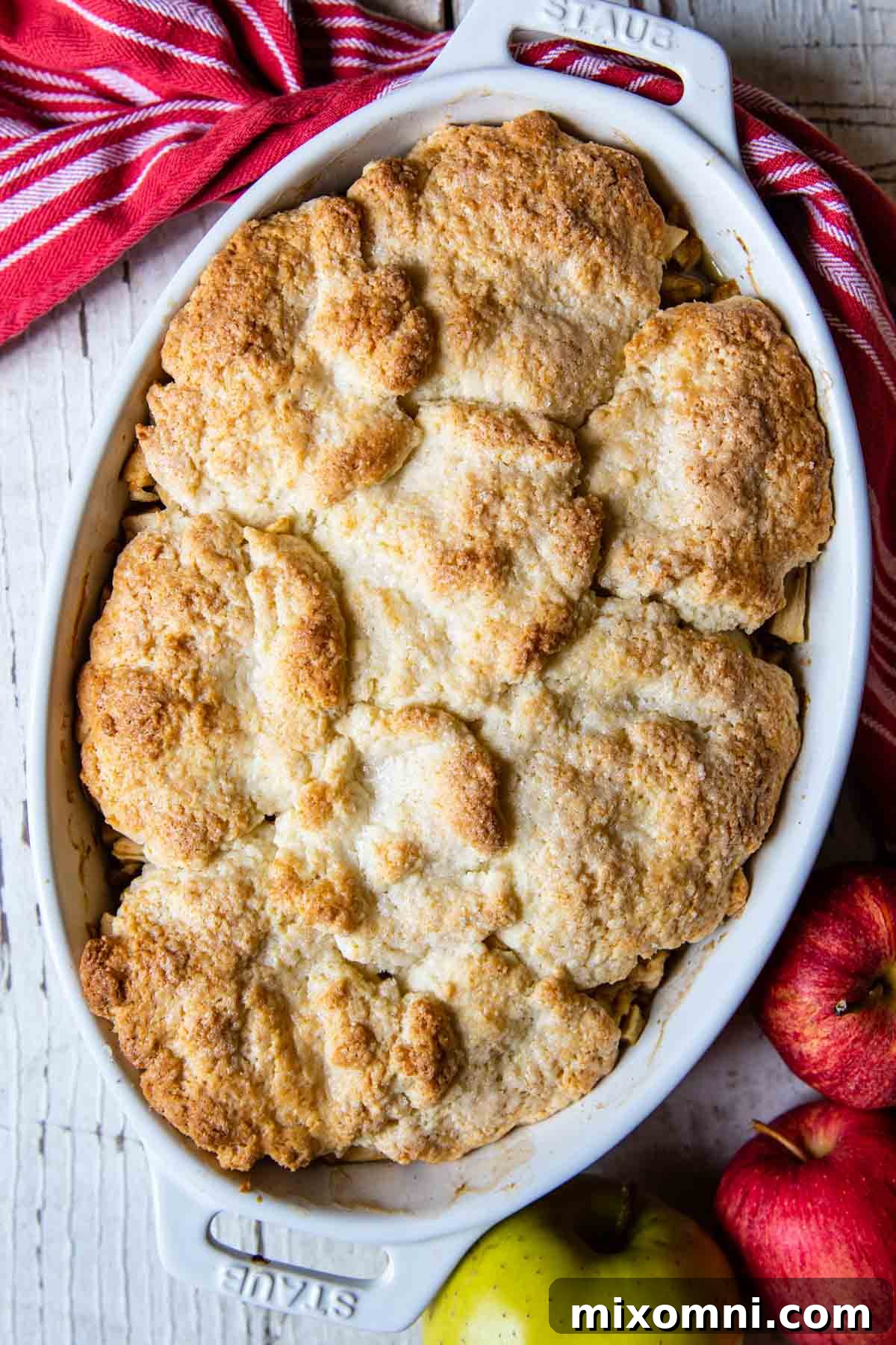 A warm gluten-free apple cobbler, beautifully golden, with fresh red and green apples scattered around and a rustic red kitchen towel, evoking a cozy autumn scene.