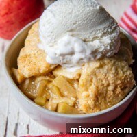 A serving of golden-brown gluten-free apple cobbler in a white bowl, with fresh apples arranged in the background.
