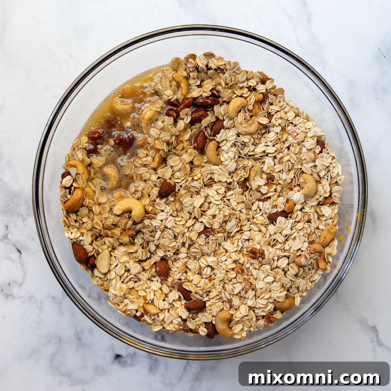 Homemade granola being mixed thoroughly with a rich maple butter mixture in a large bowl.