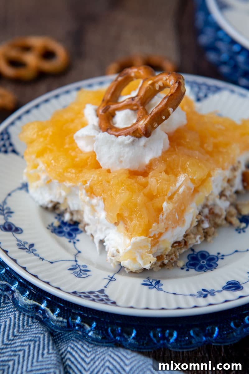 Close-up of a slice of Pineapple Pretzel Salad, showing the distinct layers of pretzel crust, cream cheese filling, and pineapple topping, garnished with whipped cream and a pretzel piece.