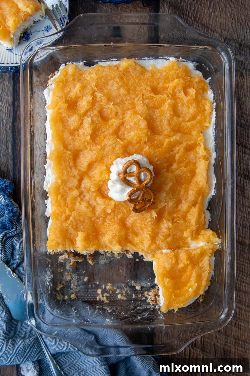 Overhead shot of Pineapple Pretzel Salad served in a clear glass baking dish, with a blue kitchen towel beside it, highlighting its vibrant colors.