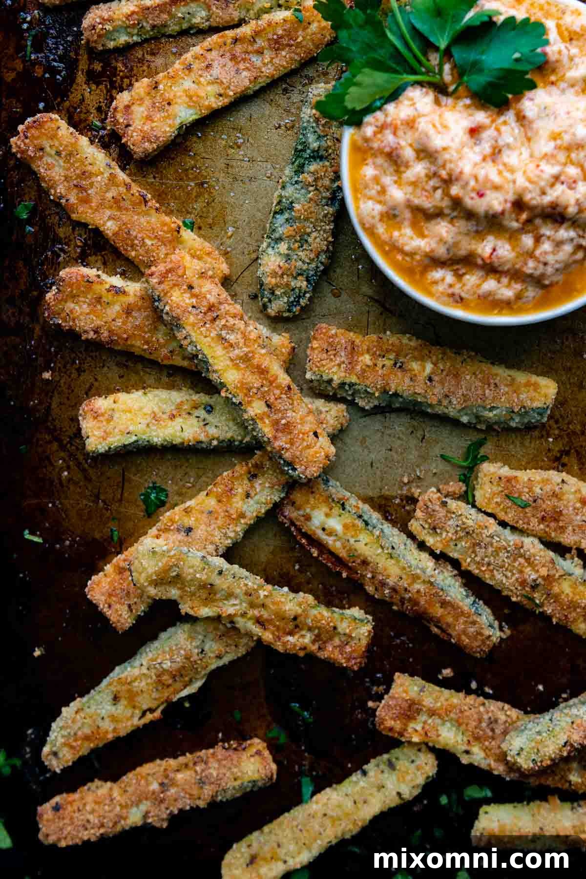 A baking sheet filled with golden, crispy baked zucchini fries, with a small bowl of sun-dried tomato aioli for dipping.