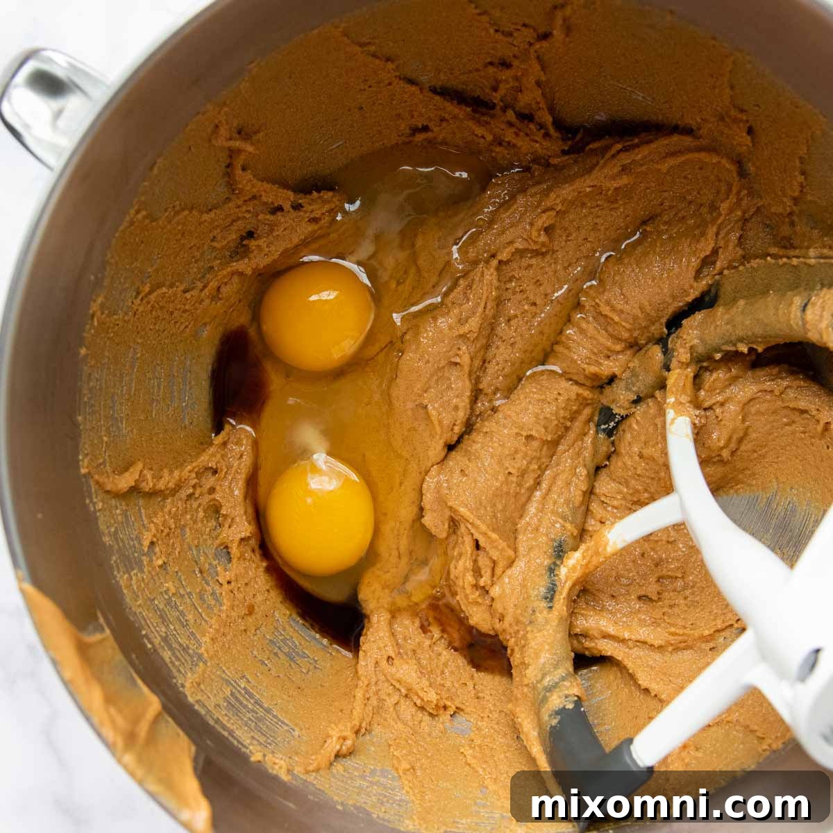 Eggs and vanilla extract being added to the creamy peanut butter and sugar mixture in a mixing bowl.