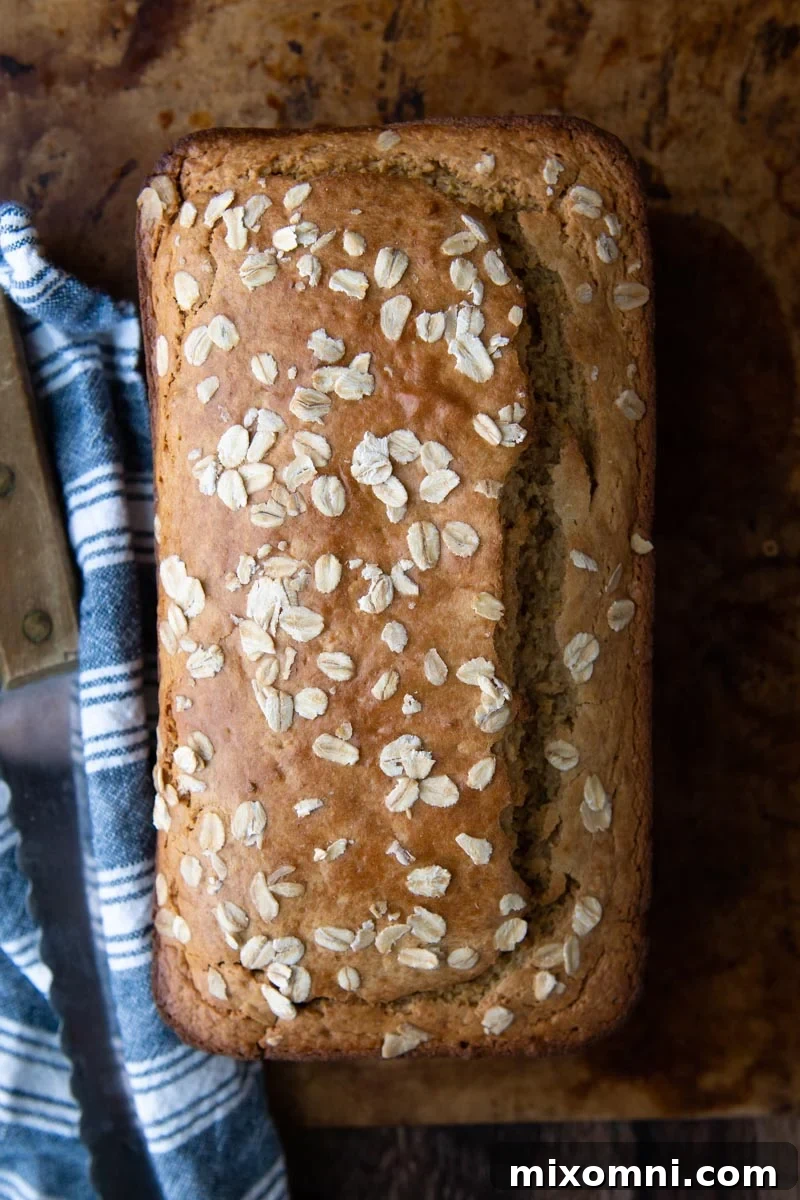 Overhead shot of a baked oat flour bread loaf, sprinkled with oats, beside a blue towel and a knife.