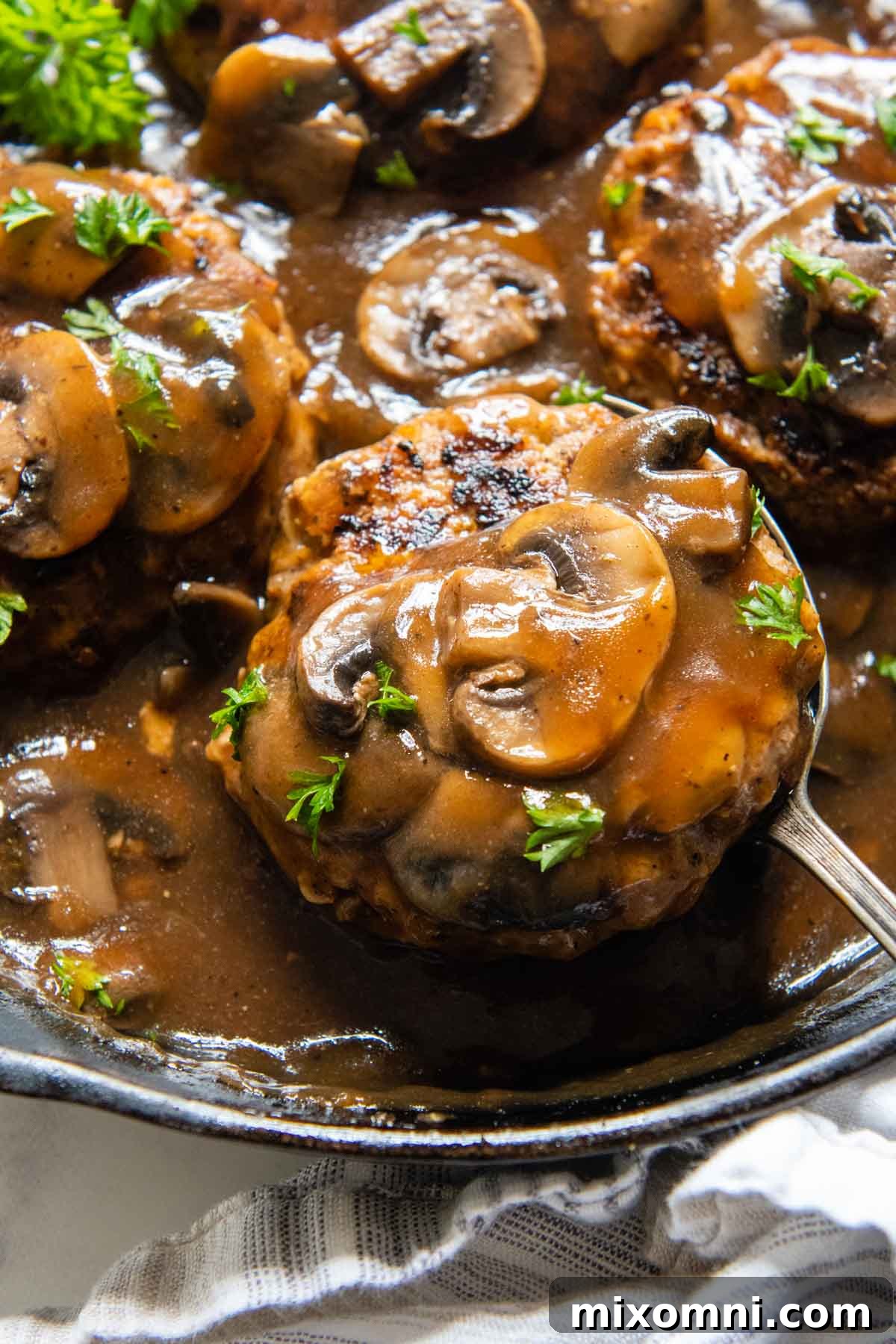 a spoon lifting up a salisbury steak with mushrooms on top from a skillet.