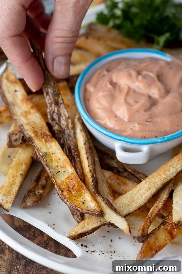 Fingers picking up a golden-brown oven baked fry from a white plate, with a small bowl of creamy fry sauce ready for dipping.