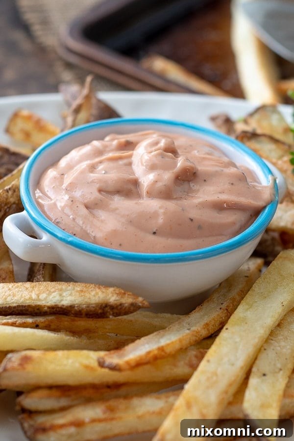 A close-up shot of a small bowl of creamy homemade fry sauce sitting on a white plate filled with golden, crispy oven baked fries, inviting a dip.