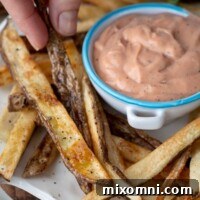 Fingers picking up an oven baked fry on a white plate with a small bowl of fry sauce